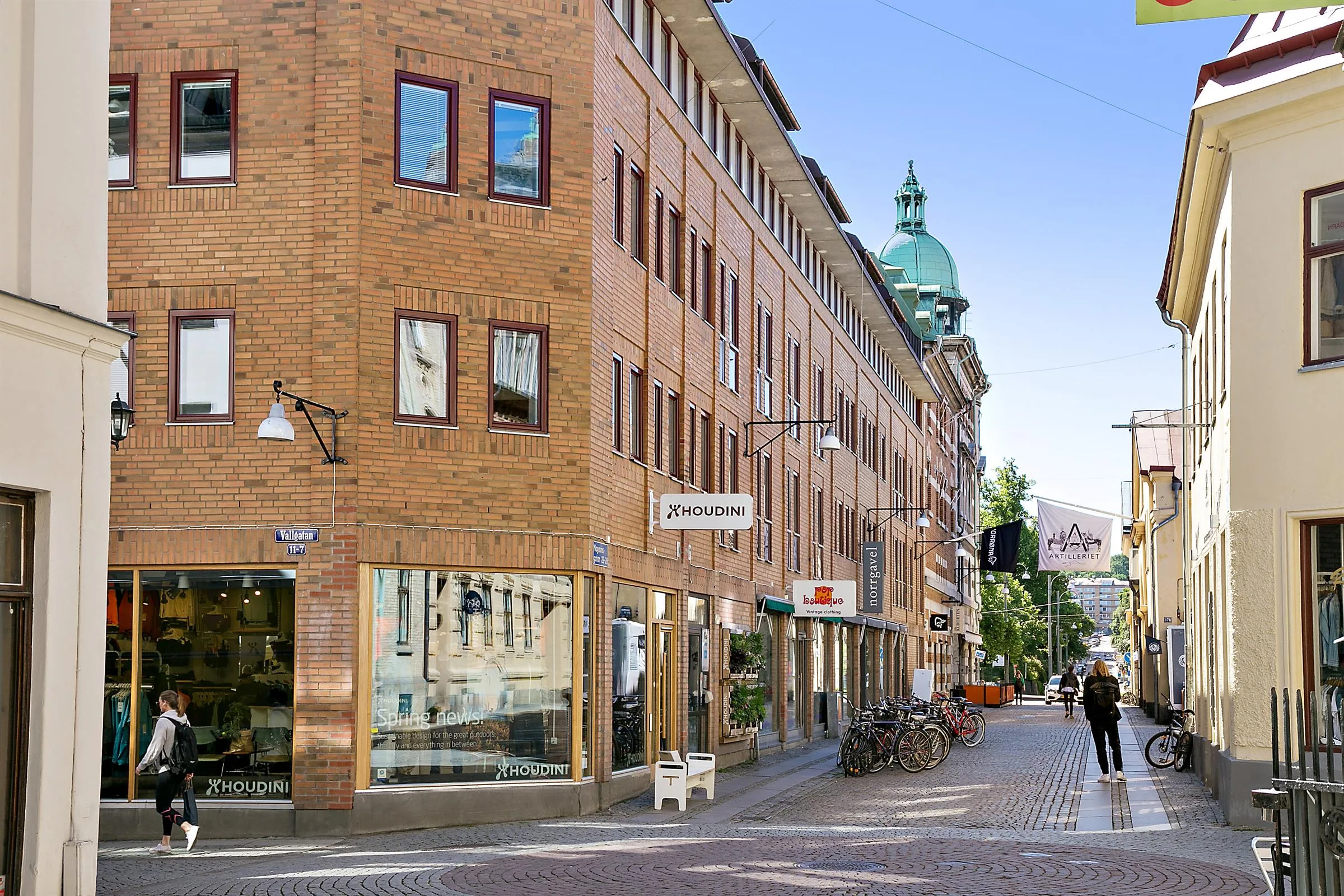 En gata med tegelbyggnader, butiker och cyklar; ett torn med grön kupol syns i bakgrunden.
