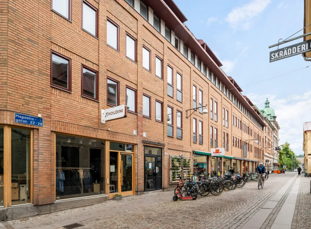 En rad tegelbyggnader med skyltar och butiksentréer, cyklar parkerade längs trottoaren.