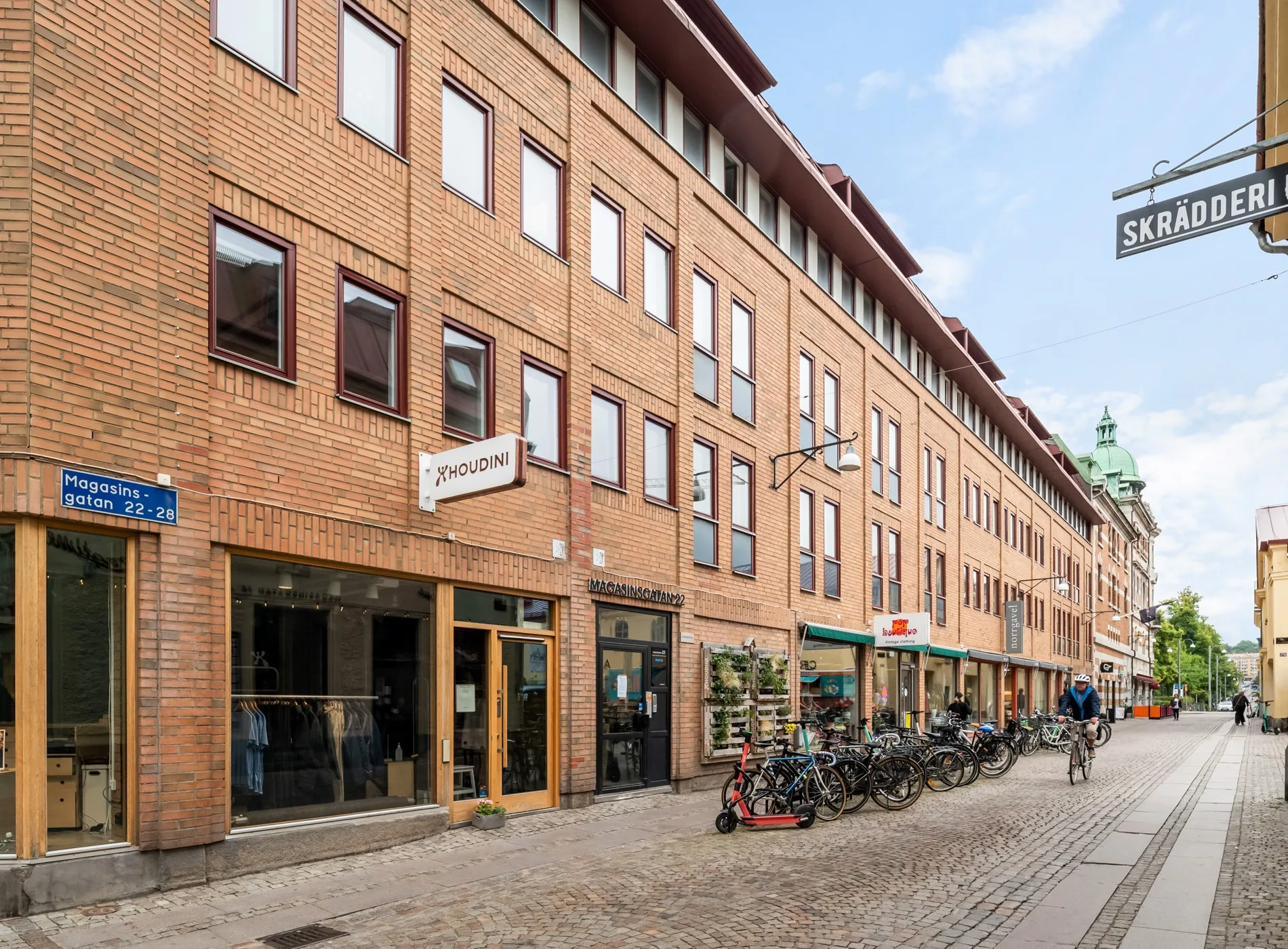 En rad tegelbyggnader med skyltar och butiksentréer, cyklar parkerade längs trottoaren.