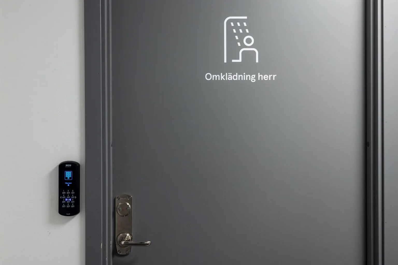 Grå dörr med texten "Omklädning herr" och en ikon av en dusch och person.