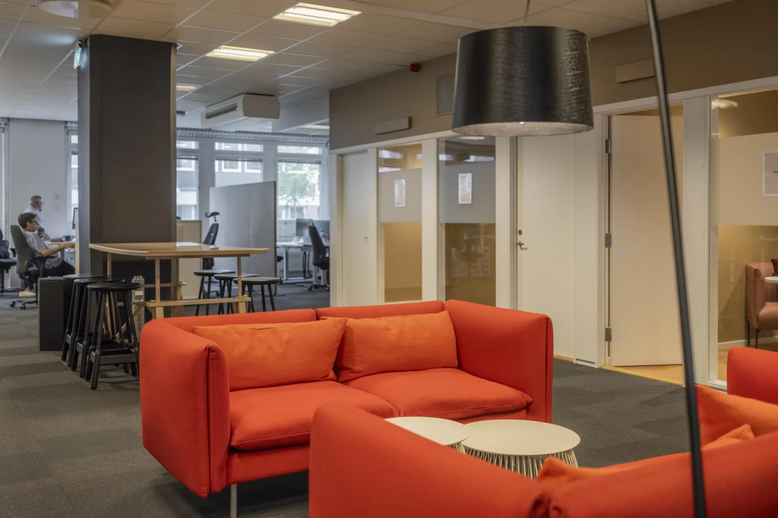 En kontorslounge med röda soffor, ett litet bord och en svart taklampa, omgiven av glasväggar och kontorsutrymmen.
