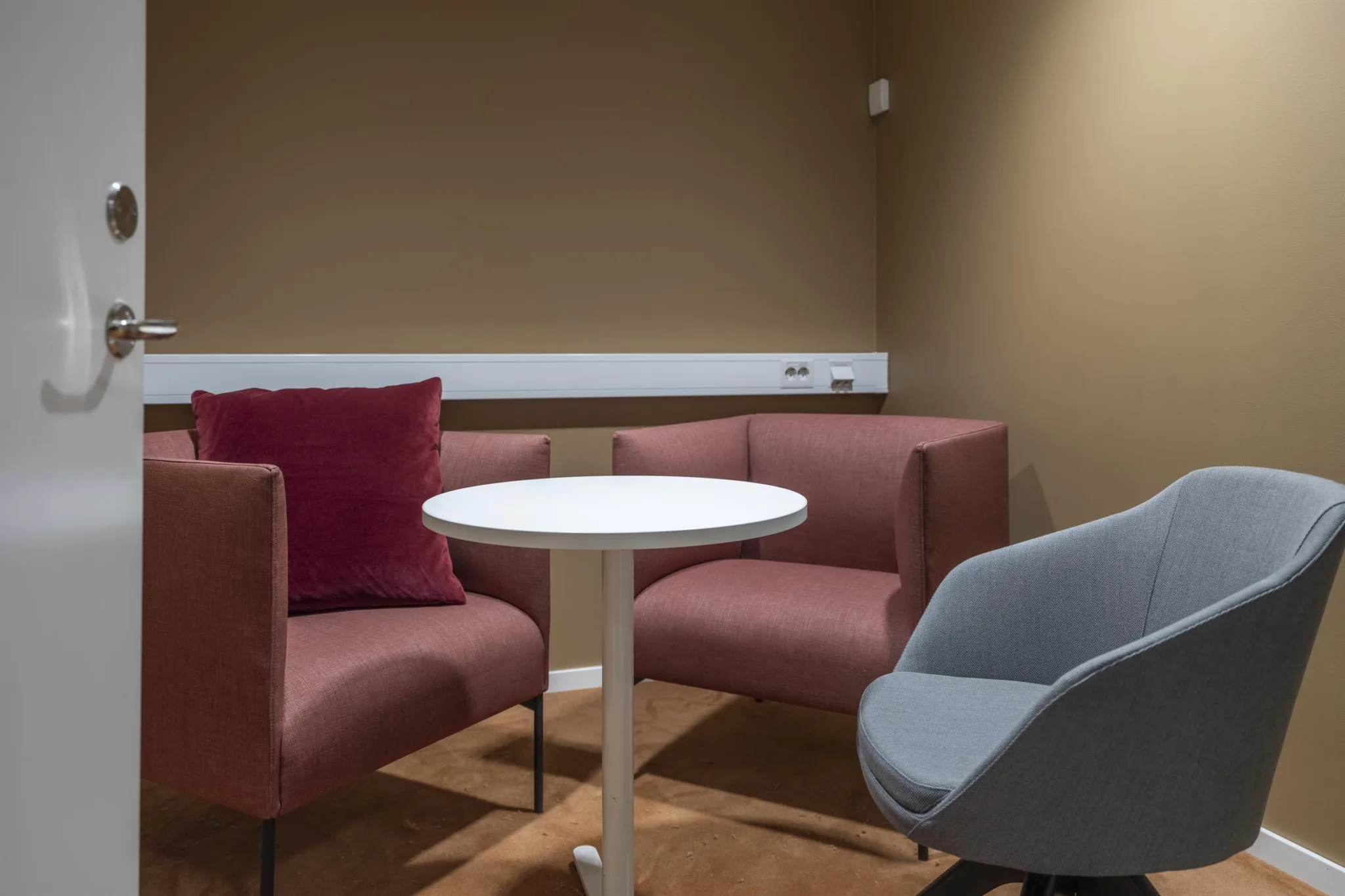 Ett litet rum med en vit rund bord, två rosa fåtöljer och en grå stol mot en beige vägg.