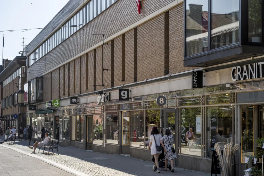 En modern byggnad med glasfasad och affärslokaler på bottenvåningen, där människor promenerar utanför.