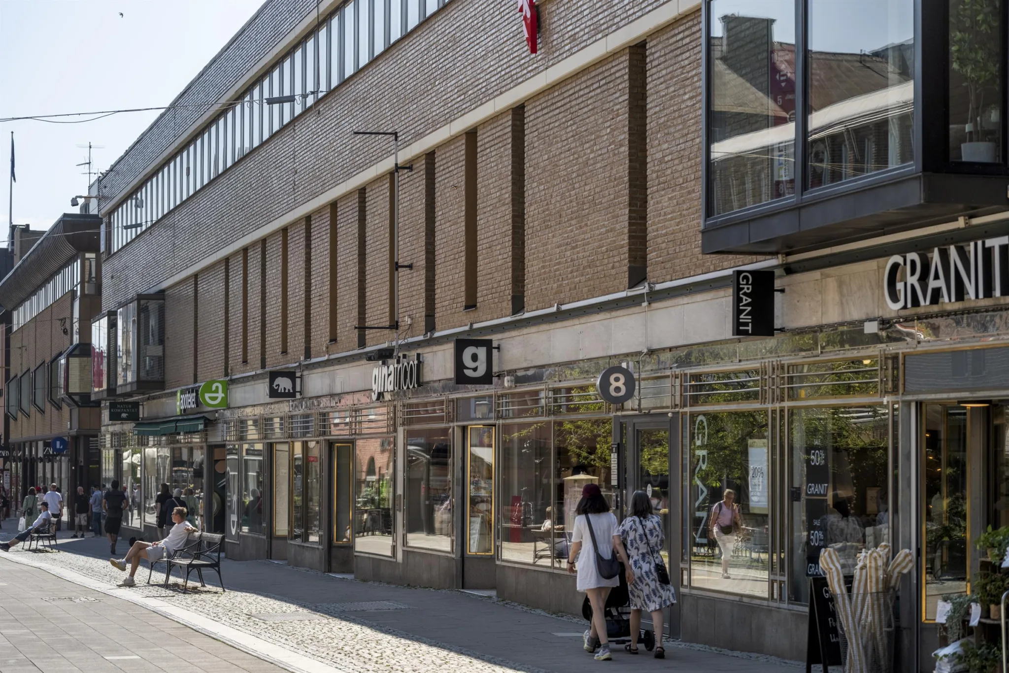En modern byggnad med glasfasad och affärslokaler på bottenvåningen, där människor promenerar utanför.
