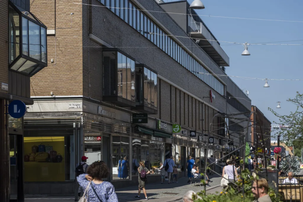 En stadsmiljö med människor som promenerar längs en gata, omgiven av byggnader och affärer.