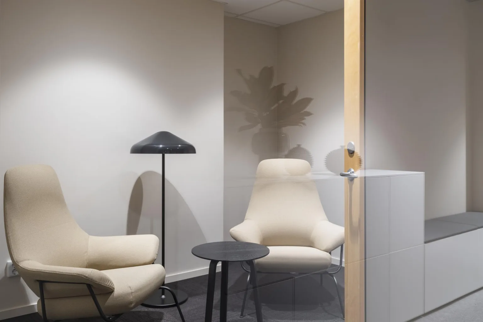 Två beige fåtöljer och ett litet bord står i ett minimalistiskt rum med en golvlampa.