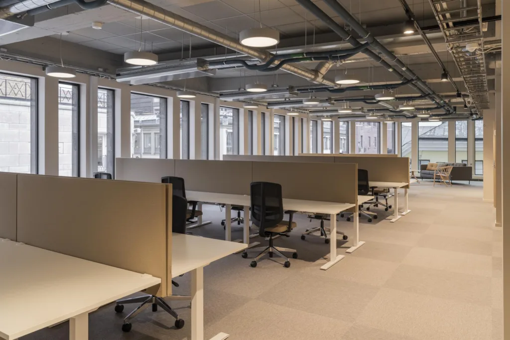 - Kontorsmiljö med flera skrivbord i rader, utrustade med kontorsstolar och skärmar, i ett rum med stora fönster och synliga ventilationsrör i taket.