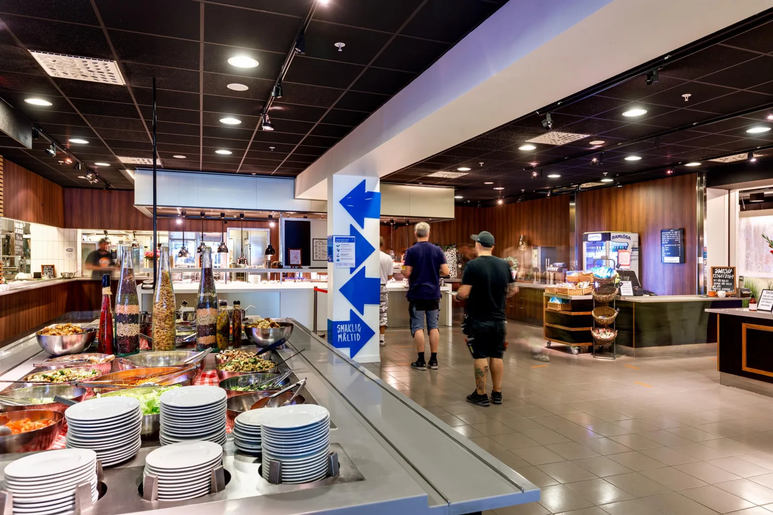 5. En restaurangmiljö med matstationer, porslin och människor som rör sig i lokalen.