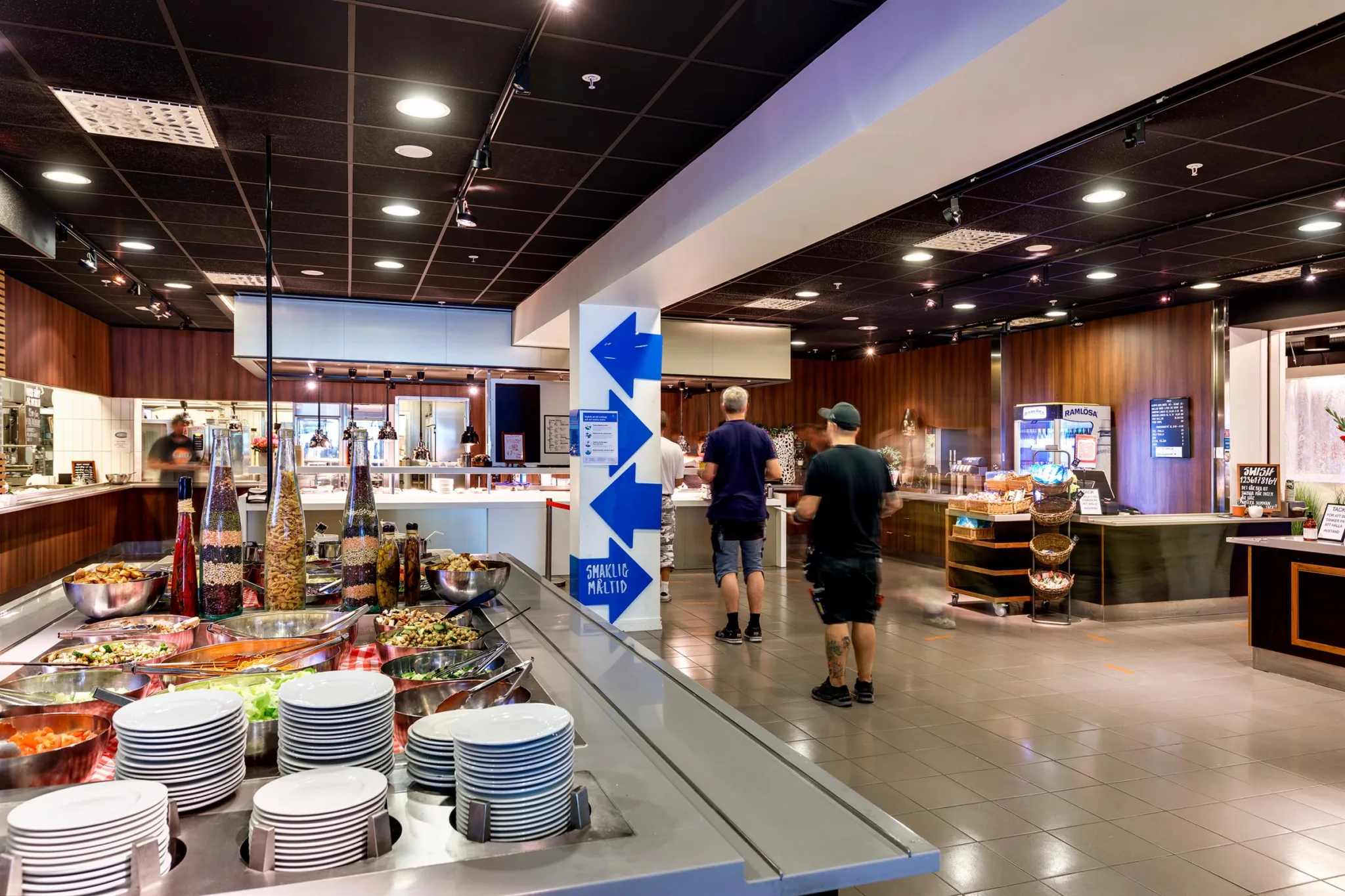 5. En restaurangmiljö med matstationer, porslin och människor som rör sig i lokalen.