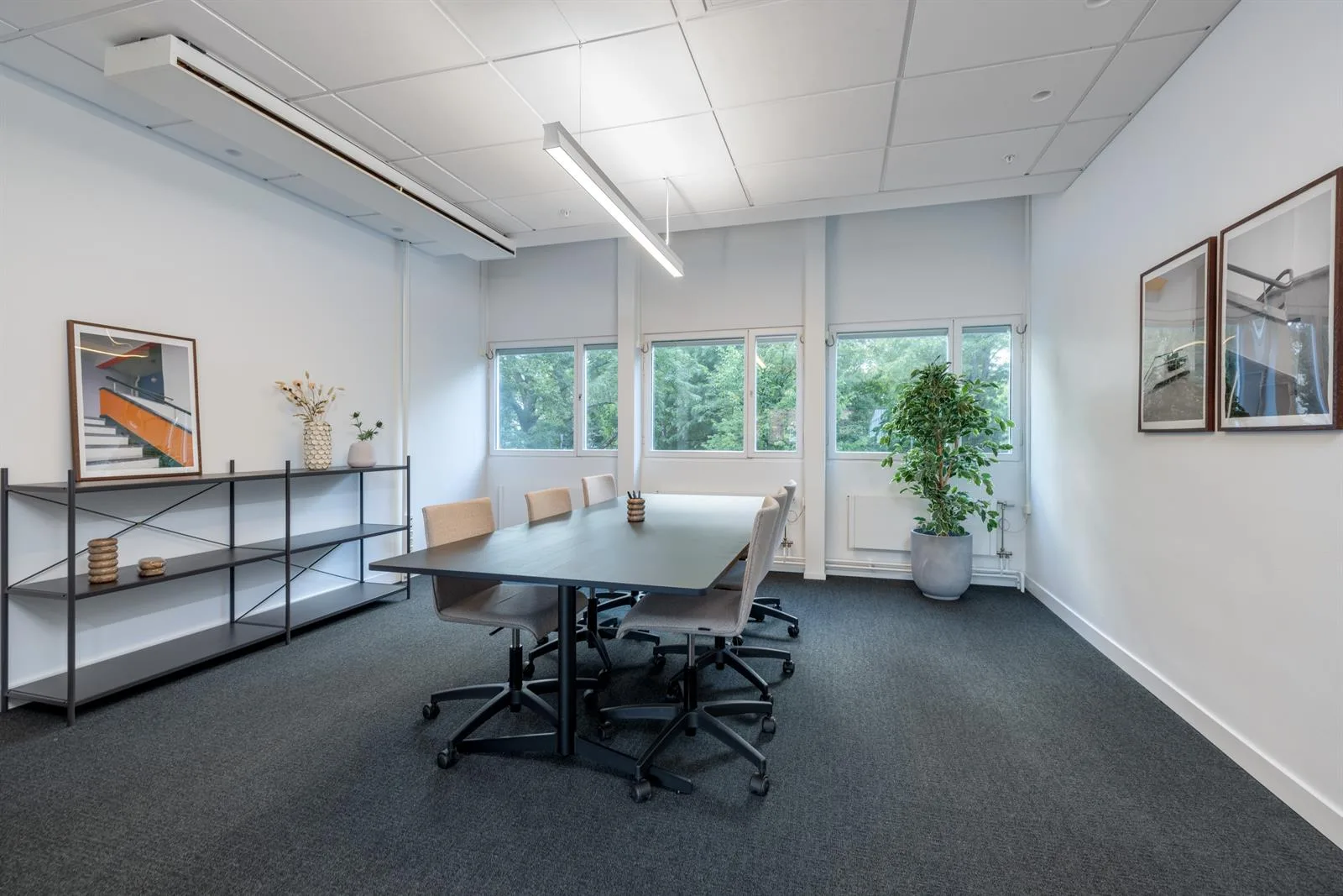 Ett ljust mötesrum med bord, stolar och hylla, stora fönster med utsikt mot grönska.