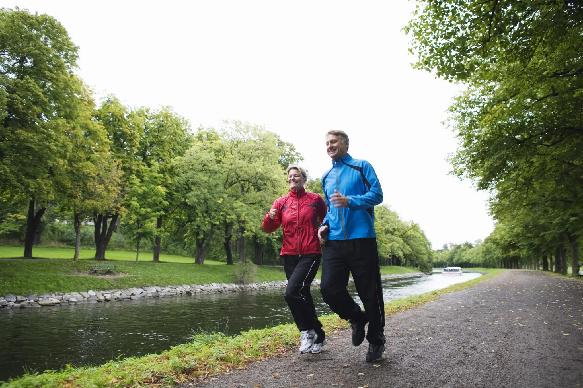 Två personer joggar längs en grusstig vid en kanal omgiven av grönskande träd.
