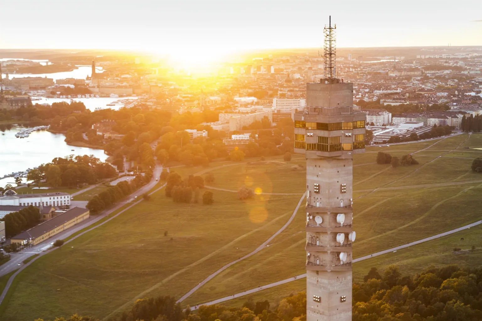 Ett högt torn med antenner, omgivet av grönområden och stadsmiljö i solnedgång.