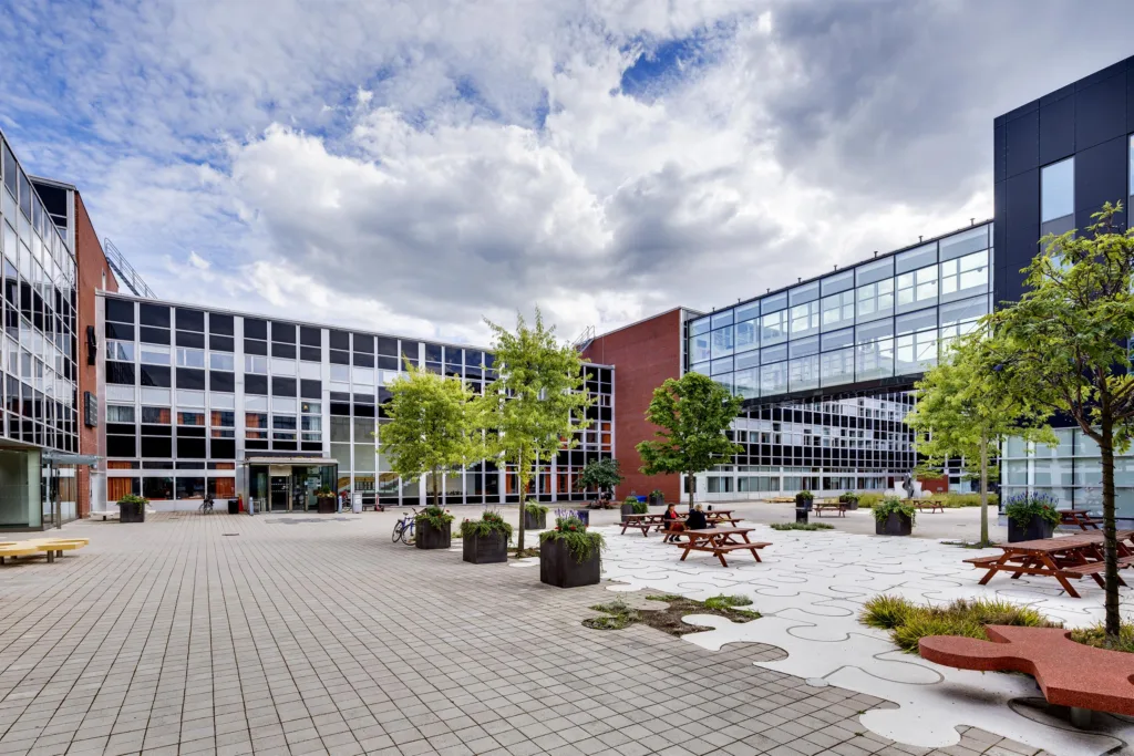 En modern byggnad med glasfasader och en innergård med träd och bänkar under en molnig himmel.