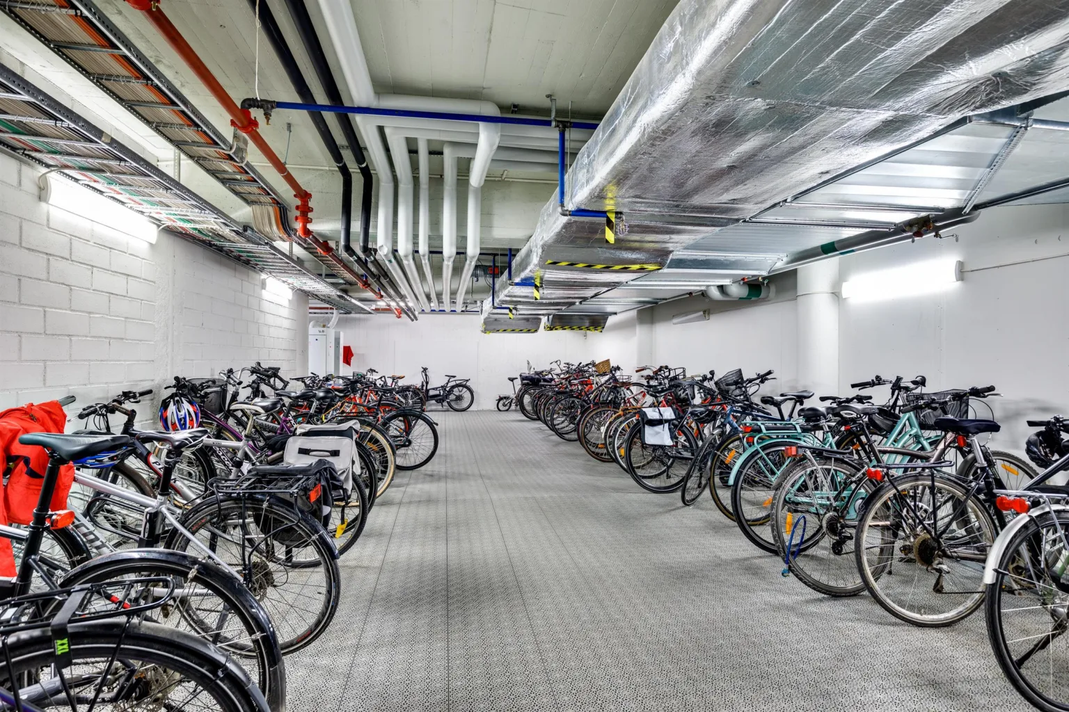 Ett cykelgarage med många cyklar parkerade i rader, rör och ventilation i taket.