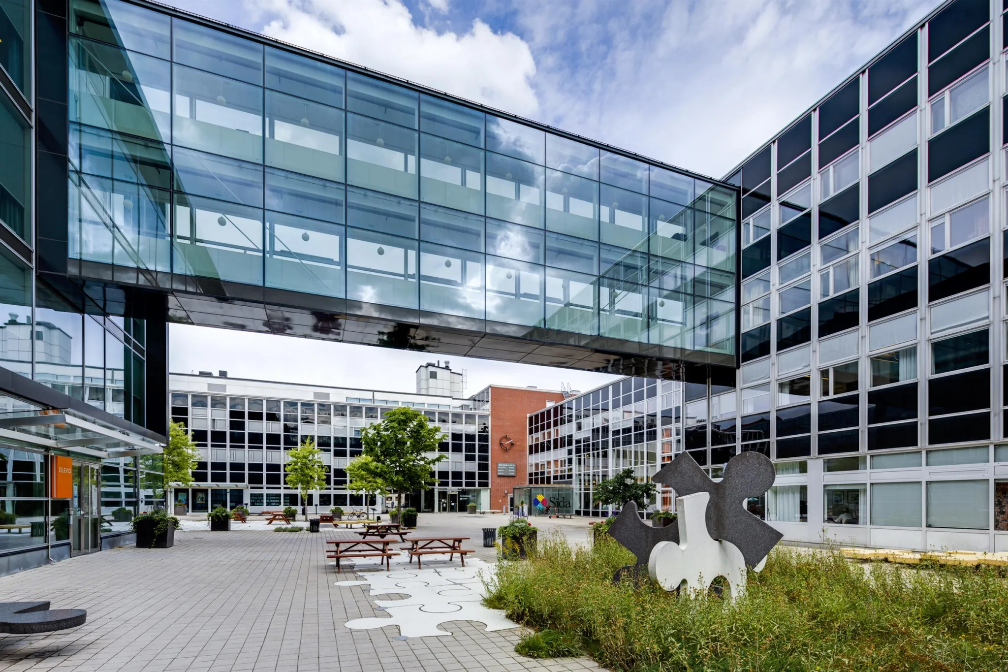 En modern byggnad med glasbro, omgiven av gröna växter och en skulptur på en gård.