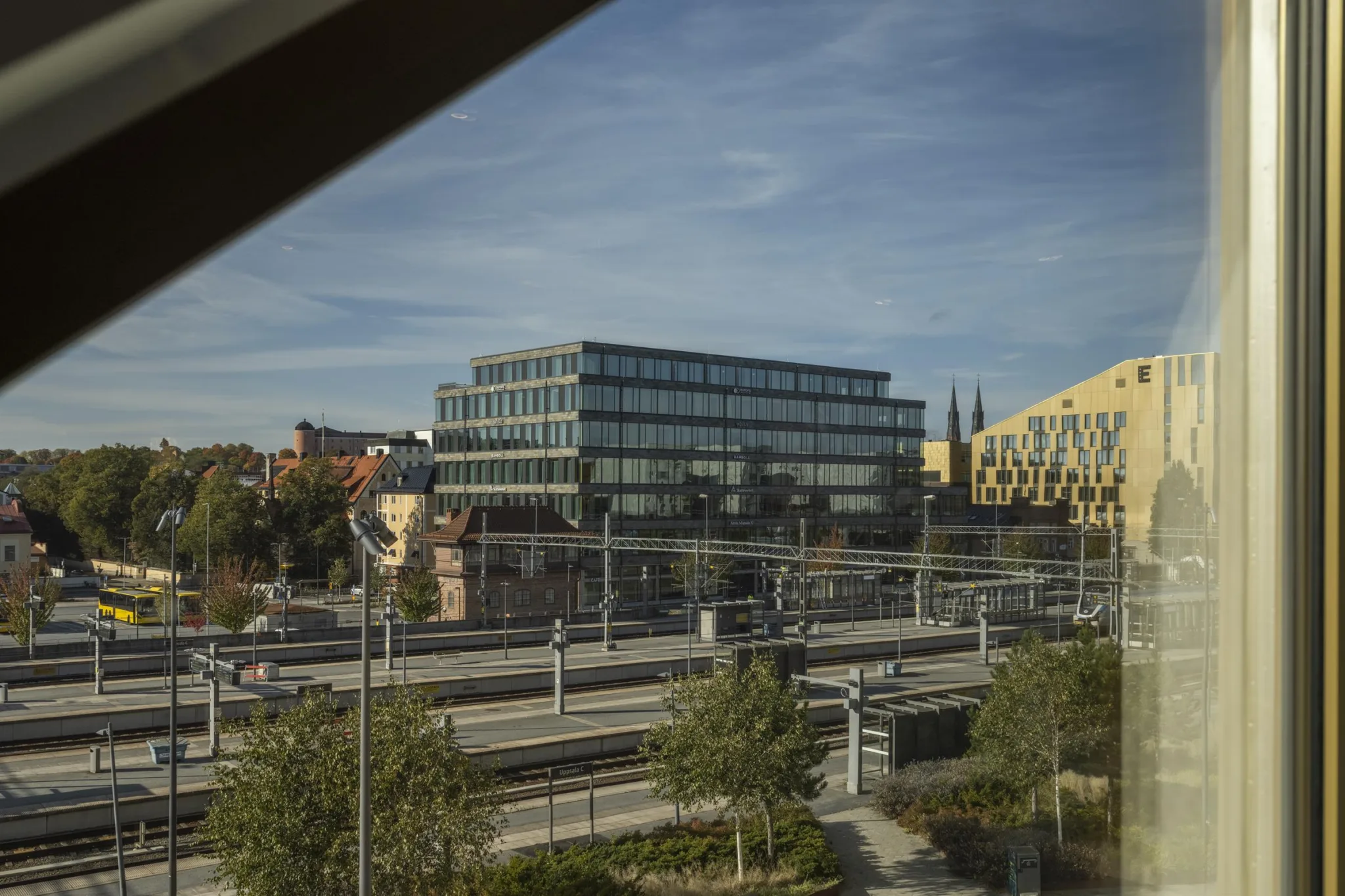 En urban utsikt med moderna byggnader, järnvägsspår och träd under en blå himmel.
