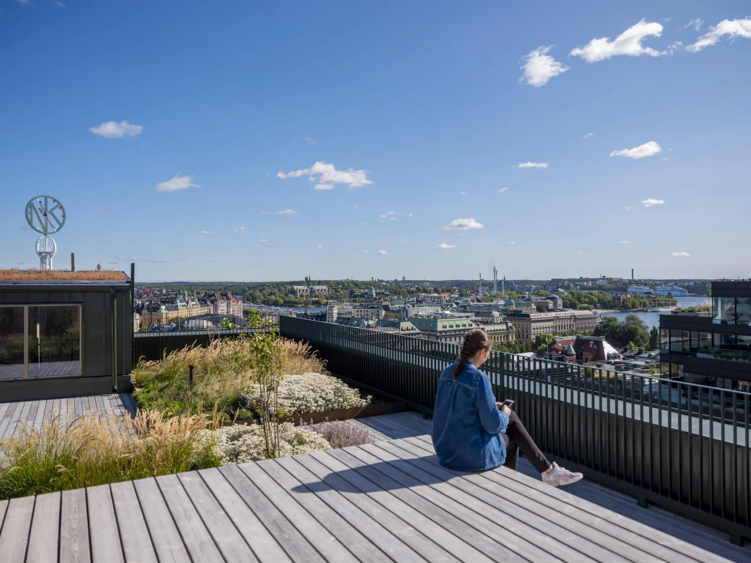 En person sitter på en träplattform på en takterrass med utsikt över en stad och blå himmel.