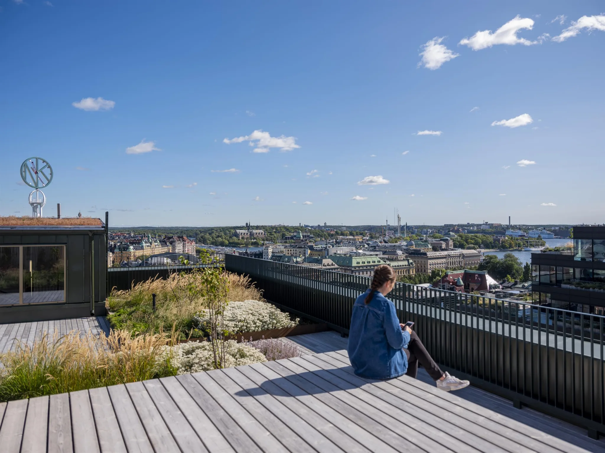 En person sitter på en träplattform på en takterrass med utsikt över en stad och blå himmel.