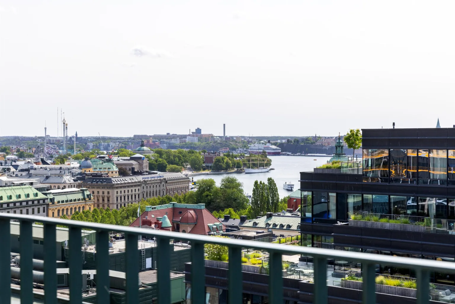 En utsikt över en stad med vatten, gröna träd och byggnader i bakgrunden, sedd från en balkong med ett grönt räcke.