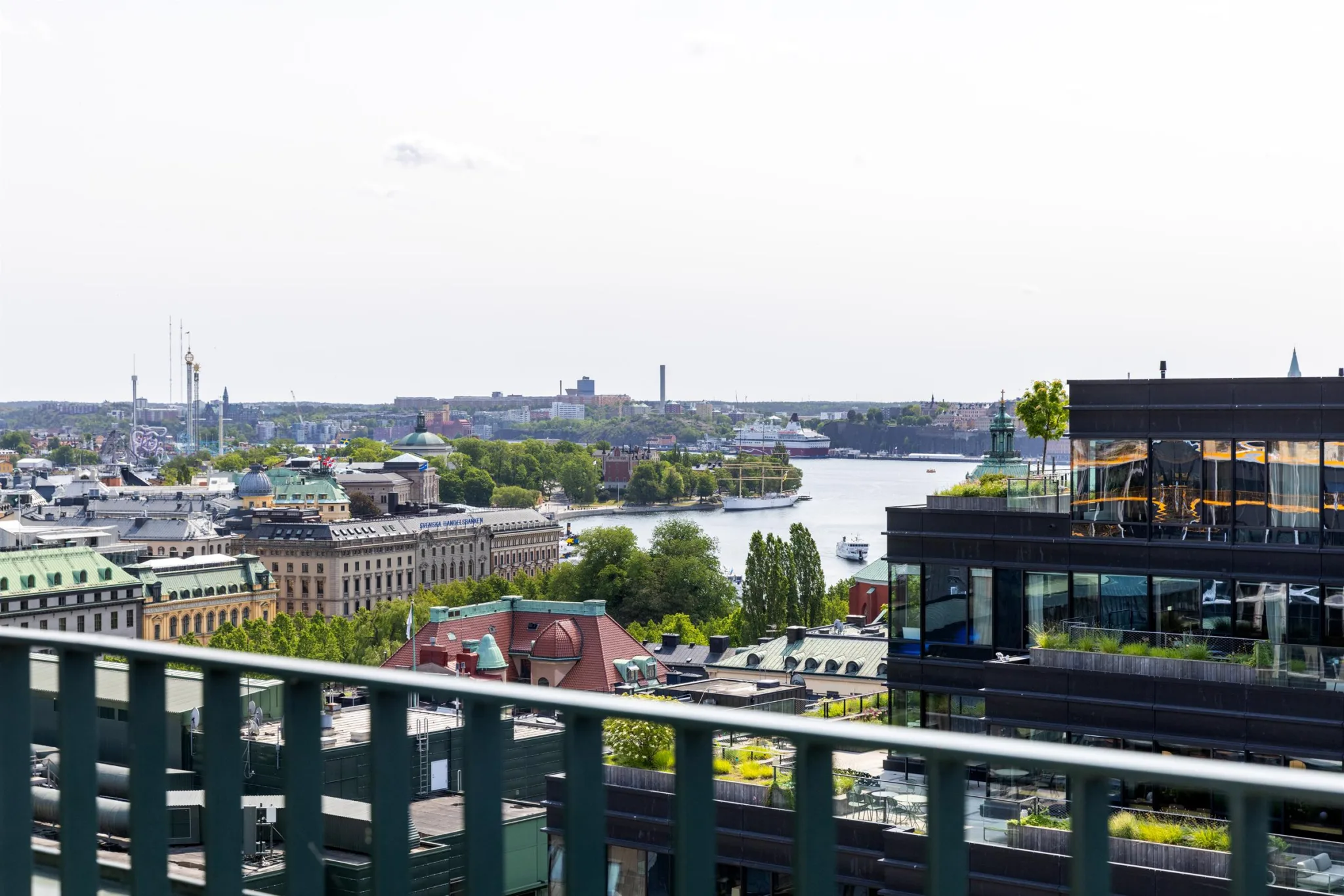En utsikt över en stad med vatten, gröna träd och byggnader i bakgrunden, sedd från en balkong med ett grönt räcke.