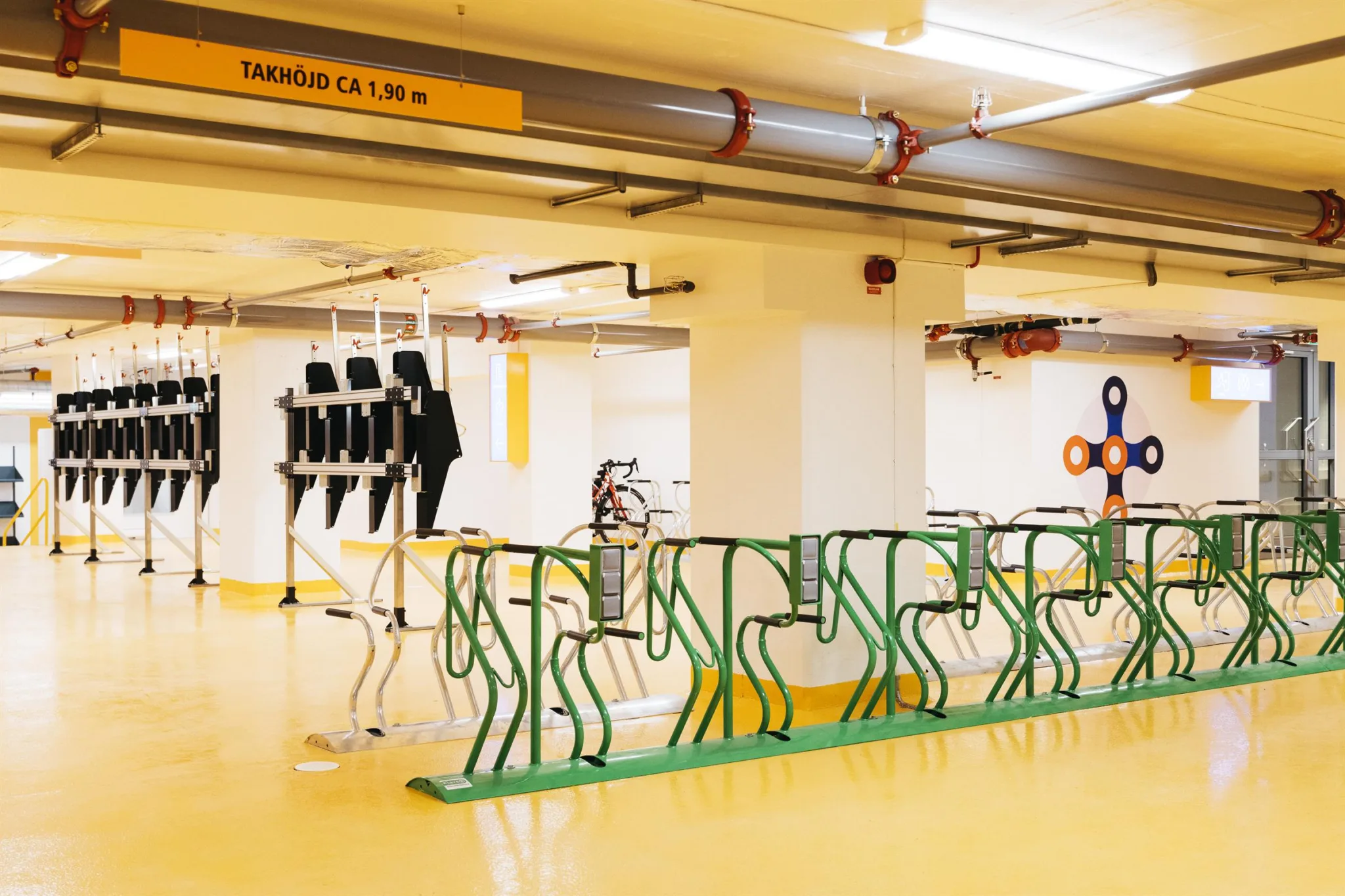En cykelparkering inomhus med gröna cykelställ, laddstationer och gulmålade golv, med en skylt som visar höjdgränsen 1,90 m.