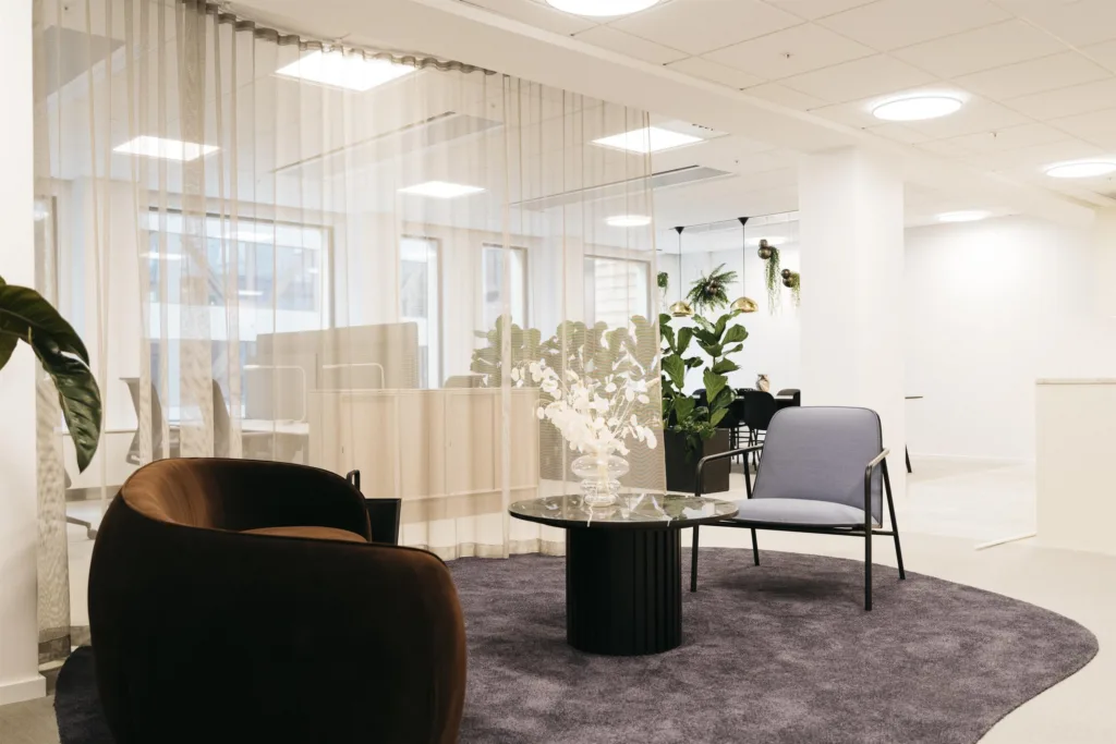 Ett modernt kontorsutrymme med sittplatser, ett runt bord, växter och glasväggar som avskiljer rummet.