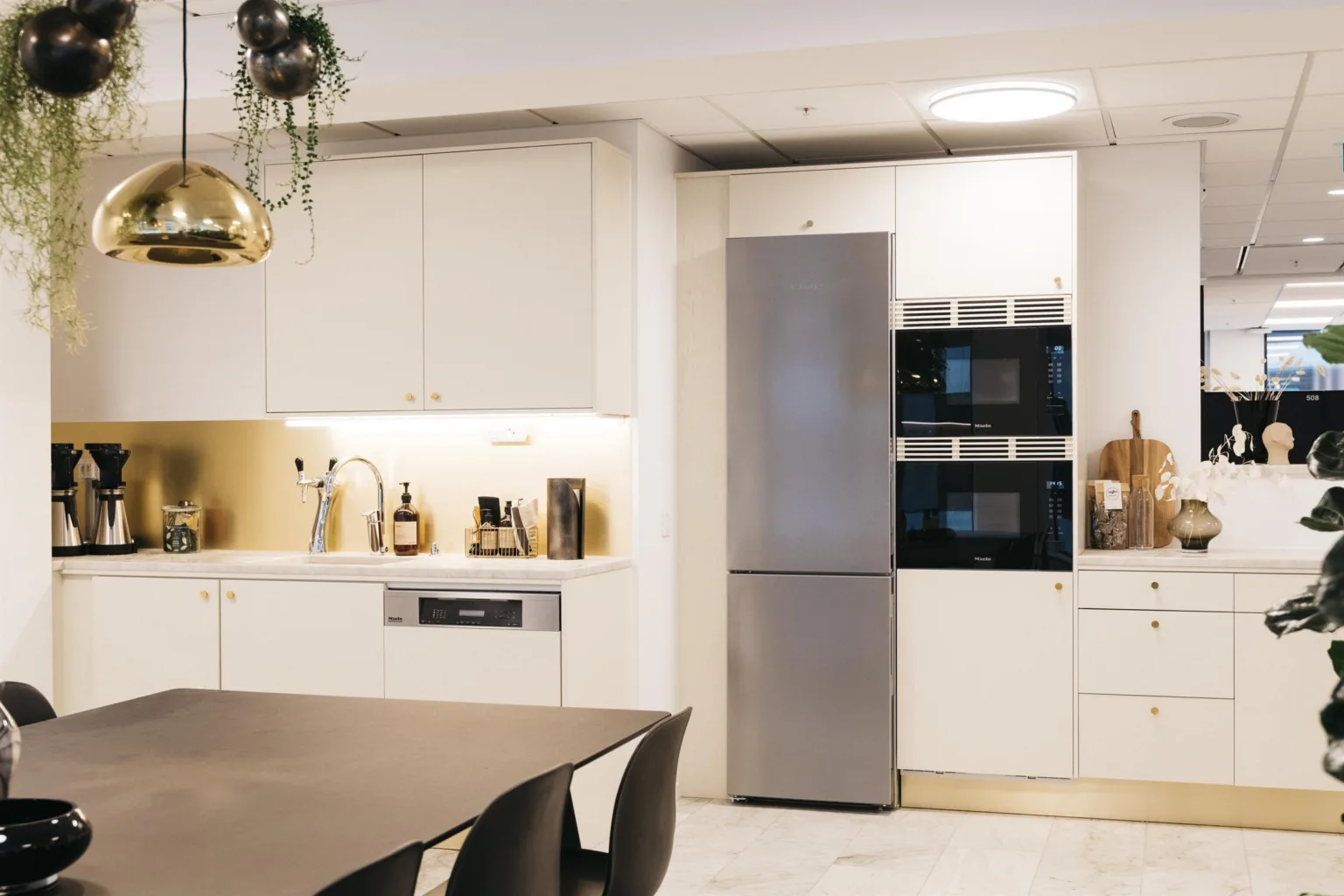 En modern köksmiljö med vita skåp, en kylskåp, inbyggda ugnar och en matplats med mörka stolar.