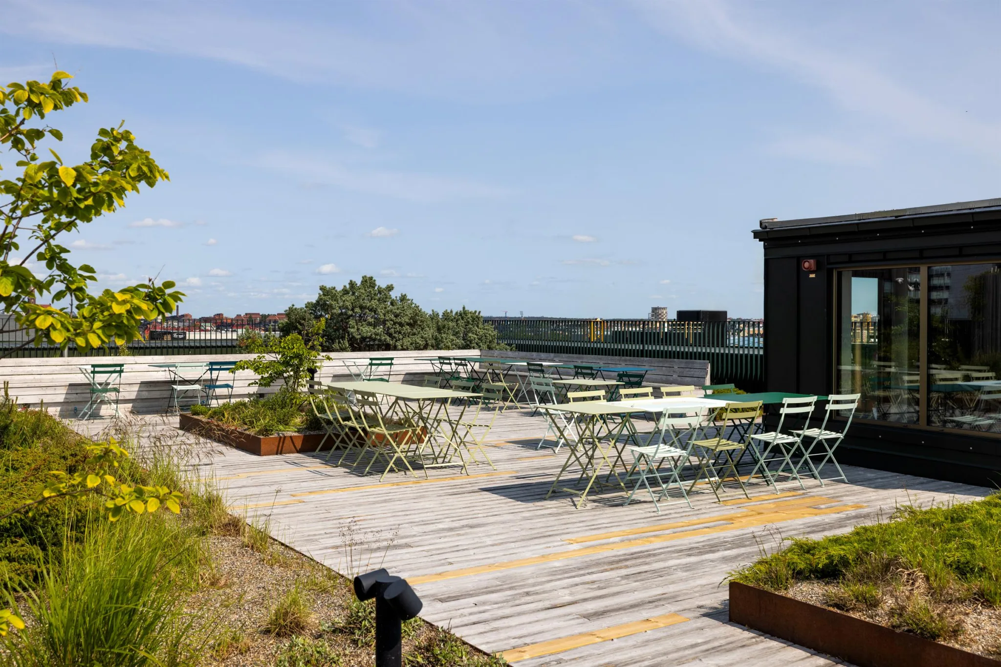 En takterrass med trätrall, bord och stolar samt grönska och utsikt över staden under en blå himmel.