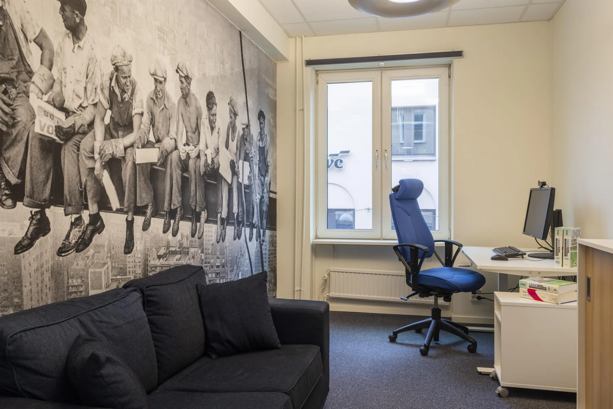 Kontorsrum med svart soffa, blå skrivbordsstol, skrivbord och en vägg med ett stort svartvitt foto.