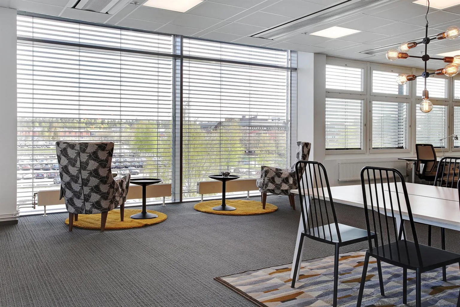 4. Loungeområde med fåtöljer, små bord och stora fönster med persienner.