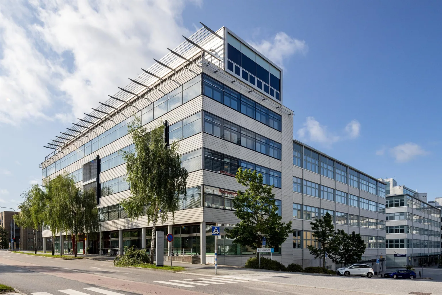 En stor kontorsbyggnad med glas och vita fasader, omgiven av träd och en trottoar.