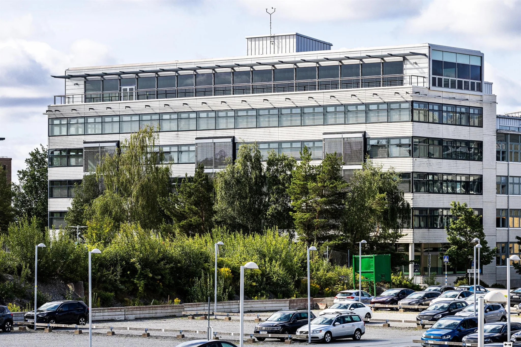 En byggnad med flera våningar, stora fönster och omgivande grönska samt parkerade bilar.