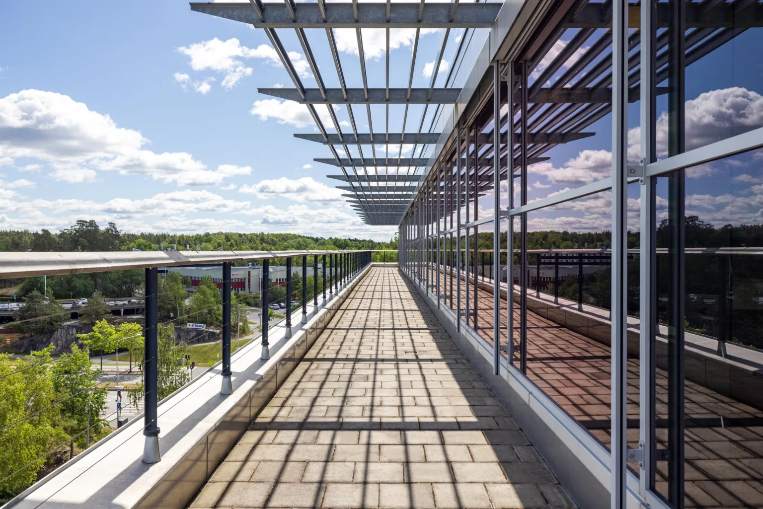 5. En lång takterrass med glasräcke och skuggor från metallstrukturen ovanför, med grön natur i bakgrunden.