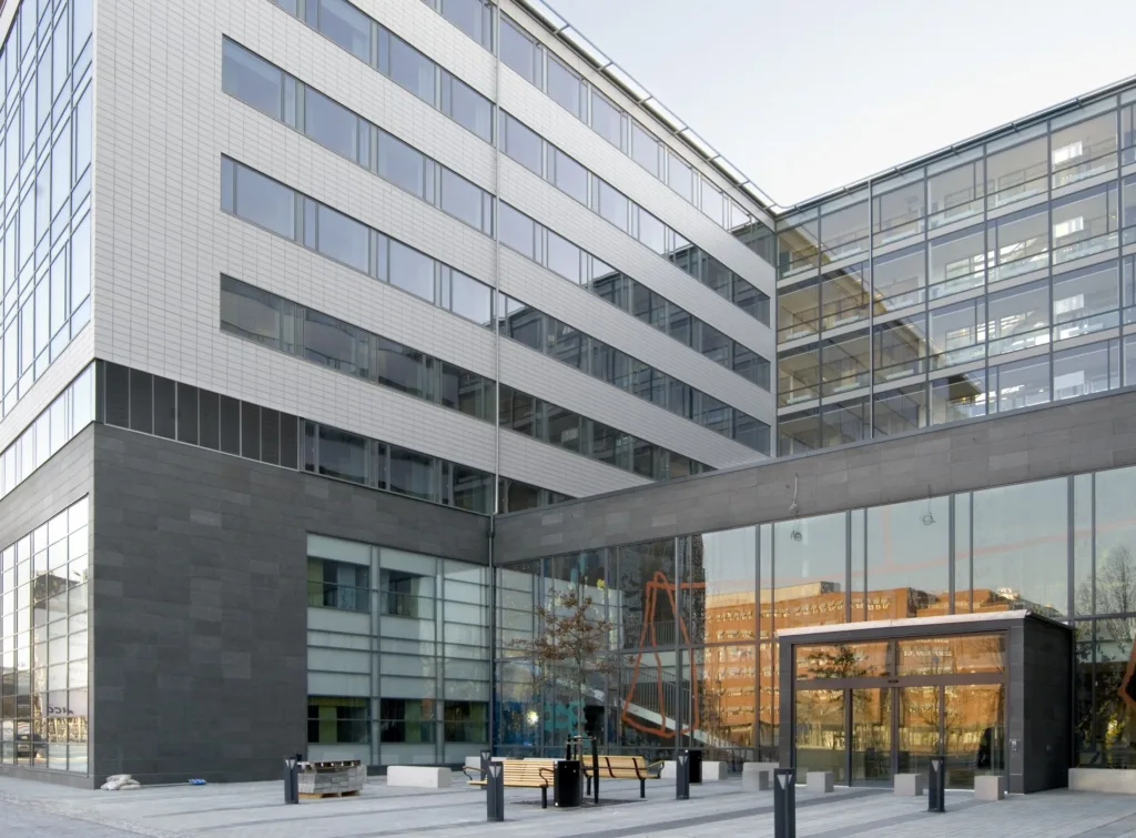 En modern kontorsbyggnad med glasfasader och flera våningar, omgiven av en gågata och bänkar.
