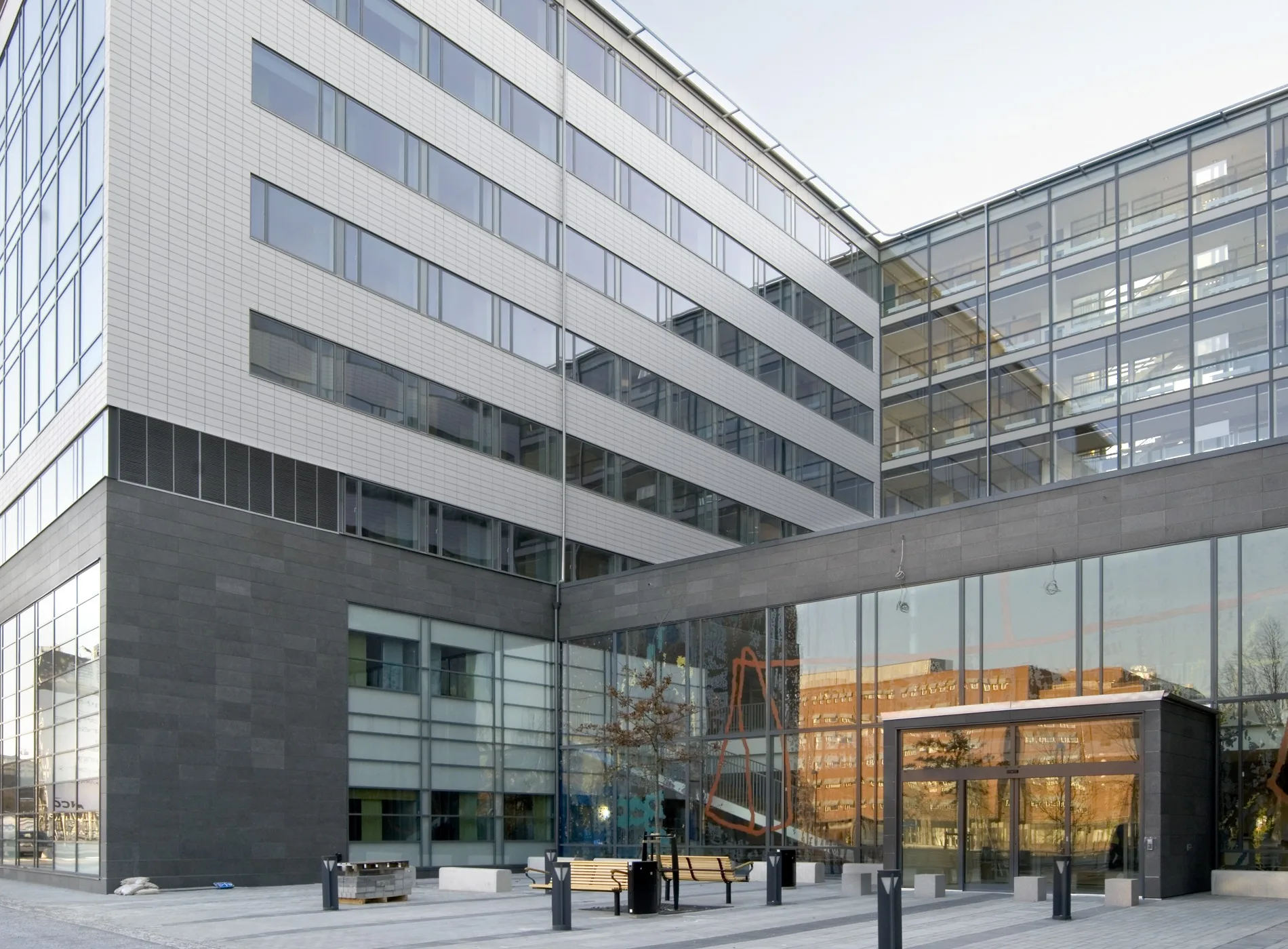 En modern kontorsbyggnad med glasfasader och flera våningar, omgiven av en gågata och bänkar.