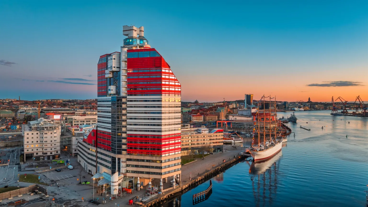 1. En modern byggnad med röd och vit fasad bredvid en hamn med en segelbåt, under en färgstark solnedgång.