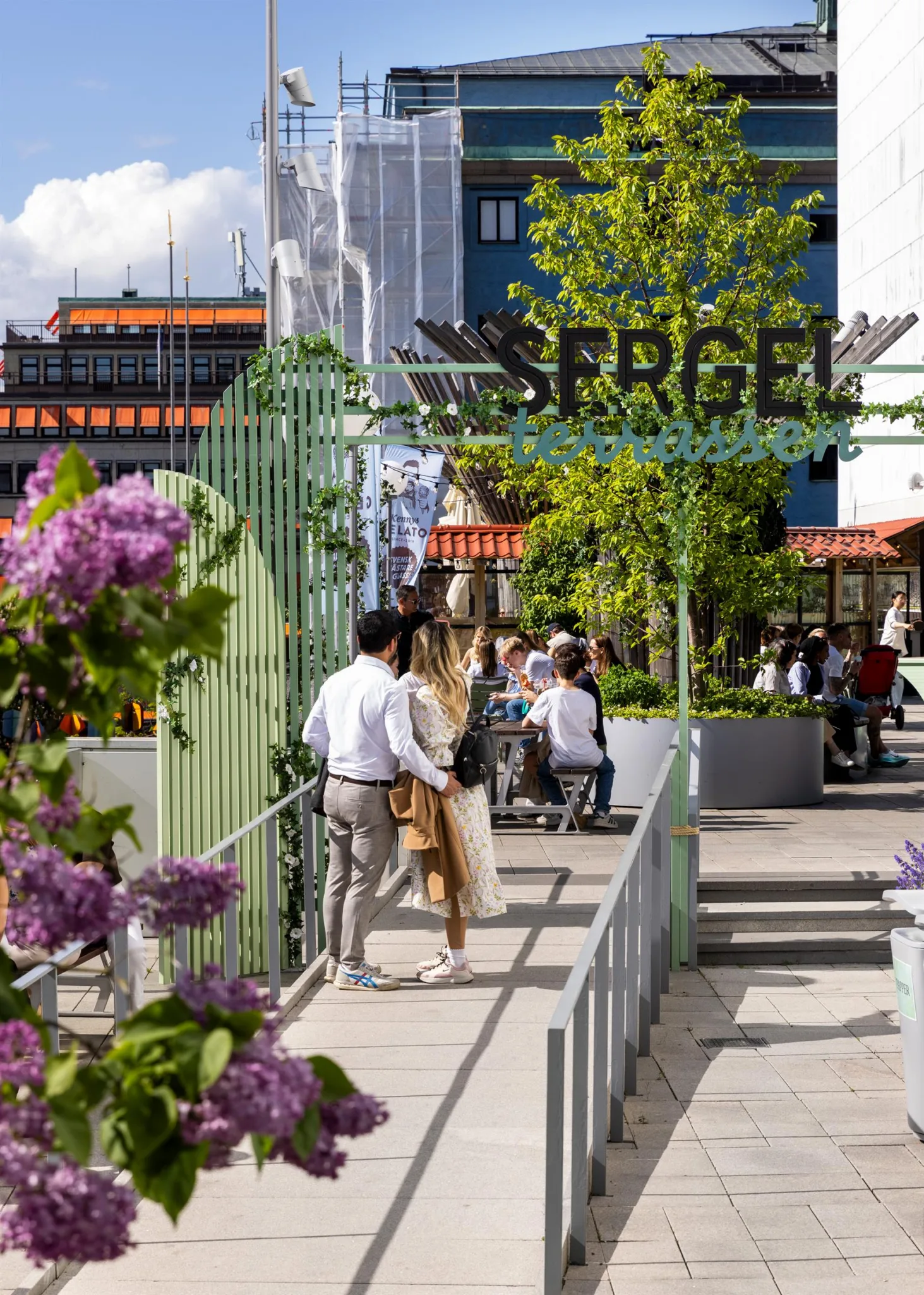 5. Grönskande takterrass med människor som promenerar och sitter, omgiven av blommor och en skylt med texten "SERGEL".