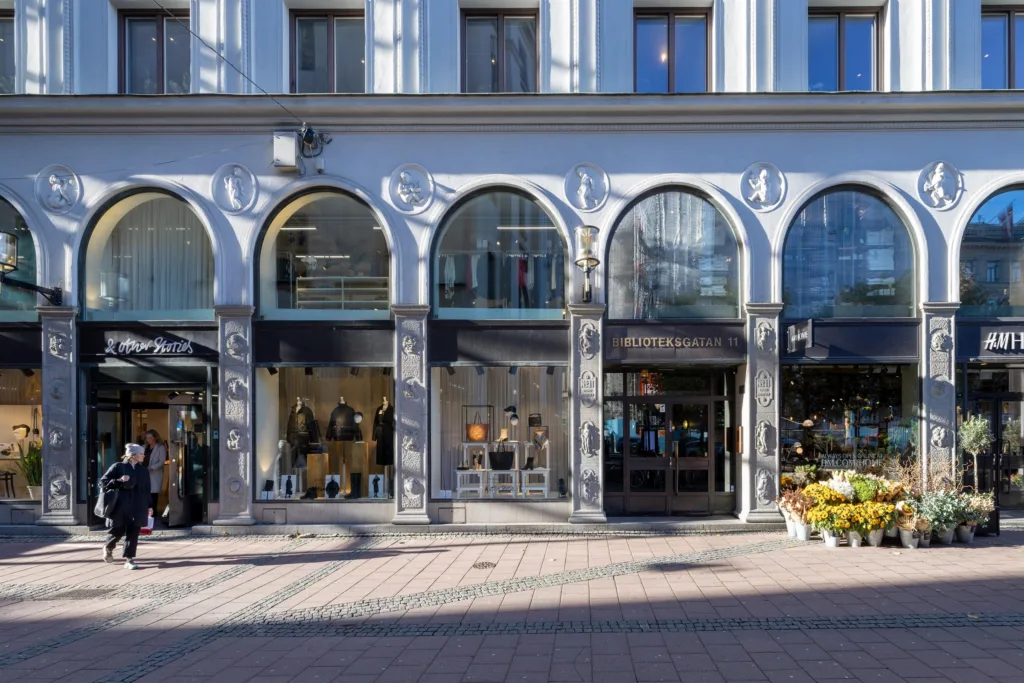 Flera butiker med välvda glasfönster i en byggnad med ljus fasad, med trottoar och skuggor i förgrunden.