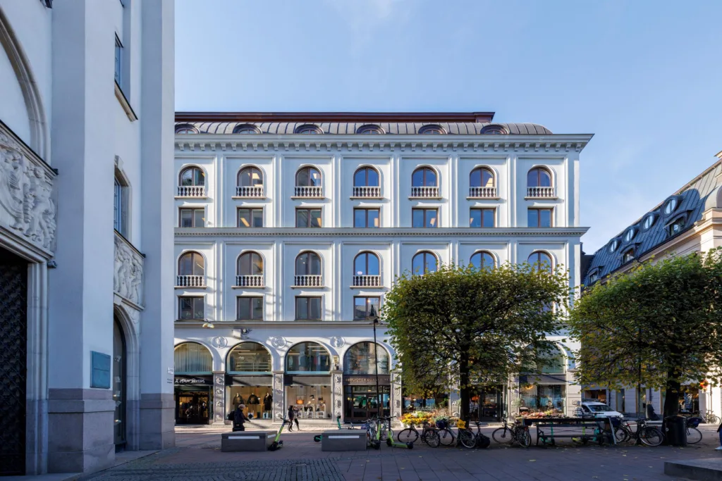 En vit byggnad med flera våningar och välvda fönster, omgiven av träd och stadsmiljö.