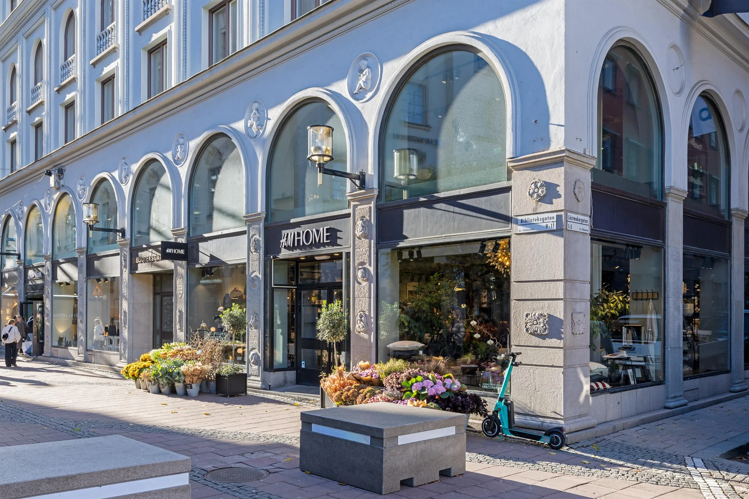 En blomsterbutik med stora välvda glasfönster, omgiven av blommor och stadsmiljö.