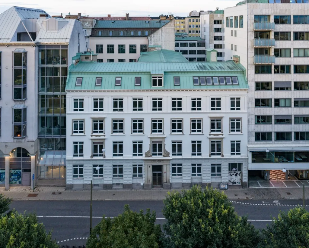 En vit byggnad med grönt tak, belägen vid en stadsgata och omgiven av moderna byggnader.