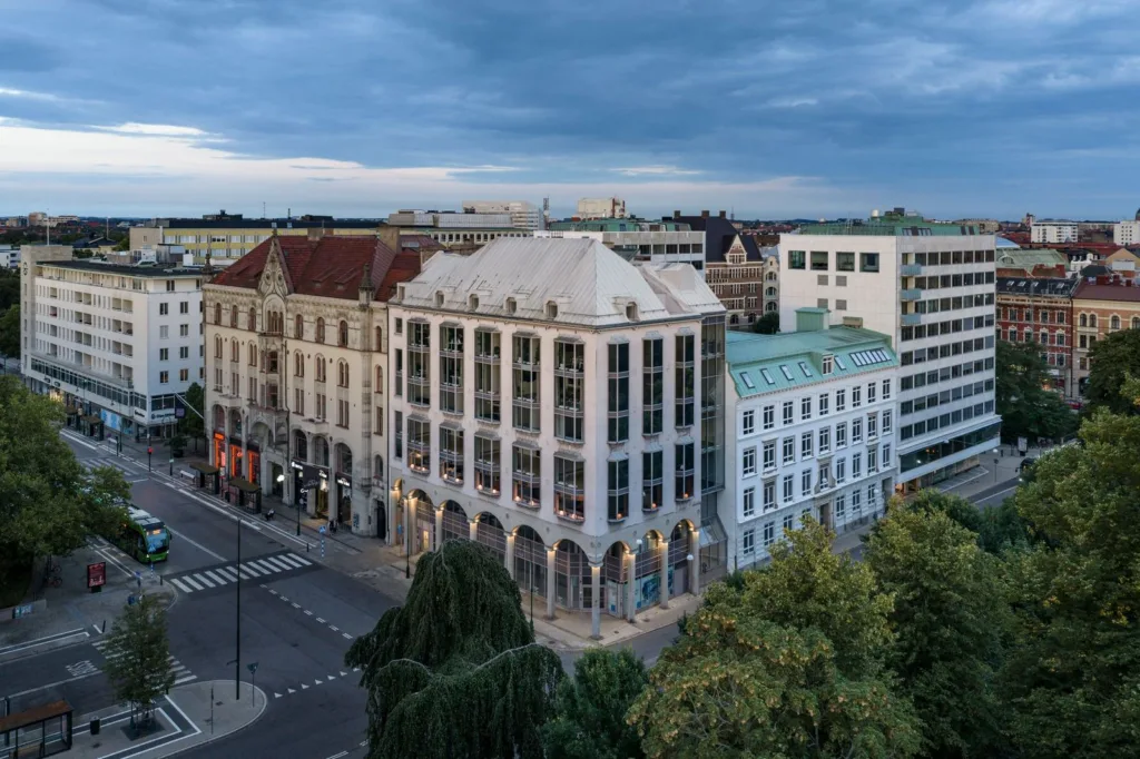 Flera historiska byggnader längs en stadsgata, omgivna av grönska och fotograferade under en molnig himmel.