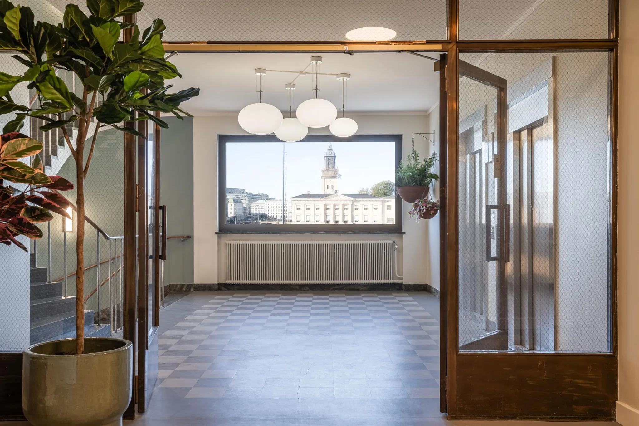 4. En inomhusgång med glasdörrar, parkettgolv, växter och stora fönster som visar en kyrka på avstånd.