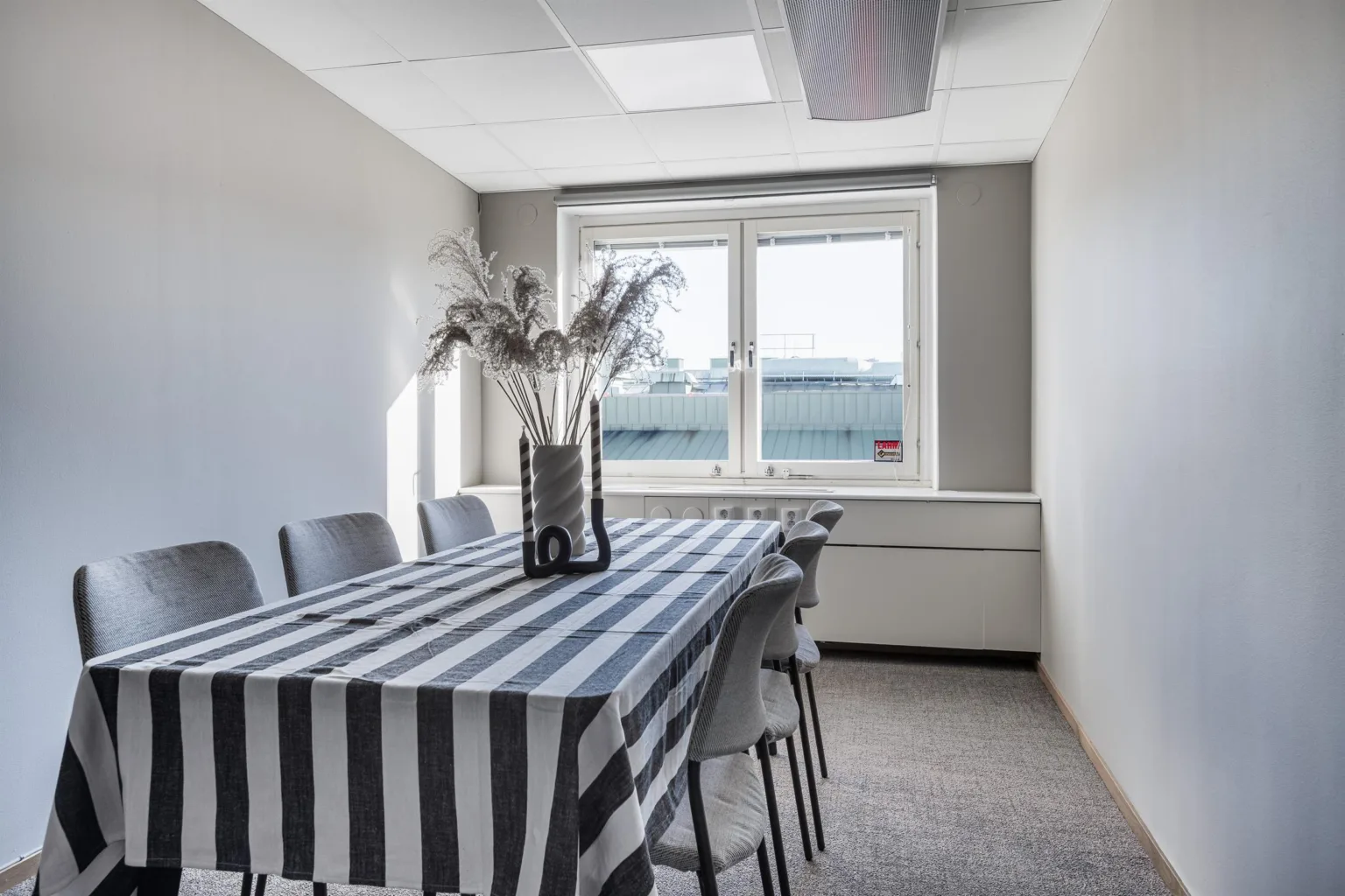 Ett konferensrum med ett bord täckt av en randig duk, omgiven av stolar och dekorerat med en vas med torkade växter, mot ett fönster.