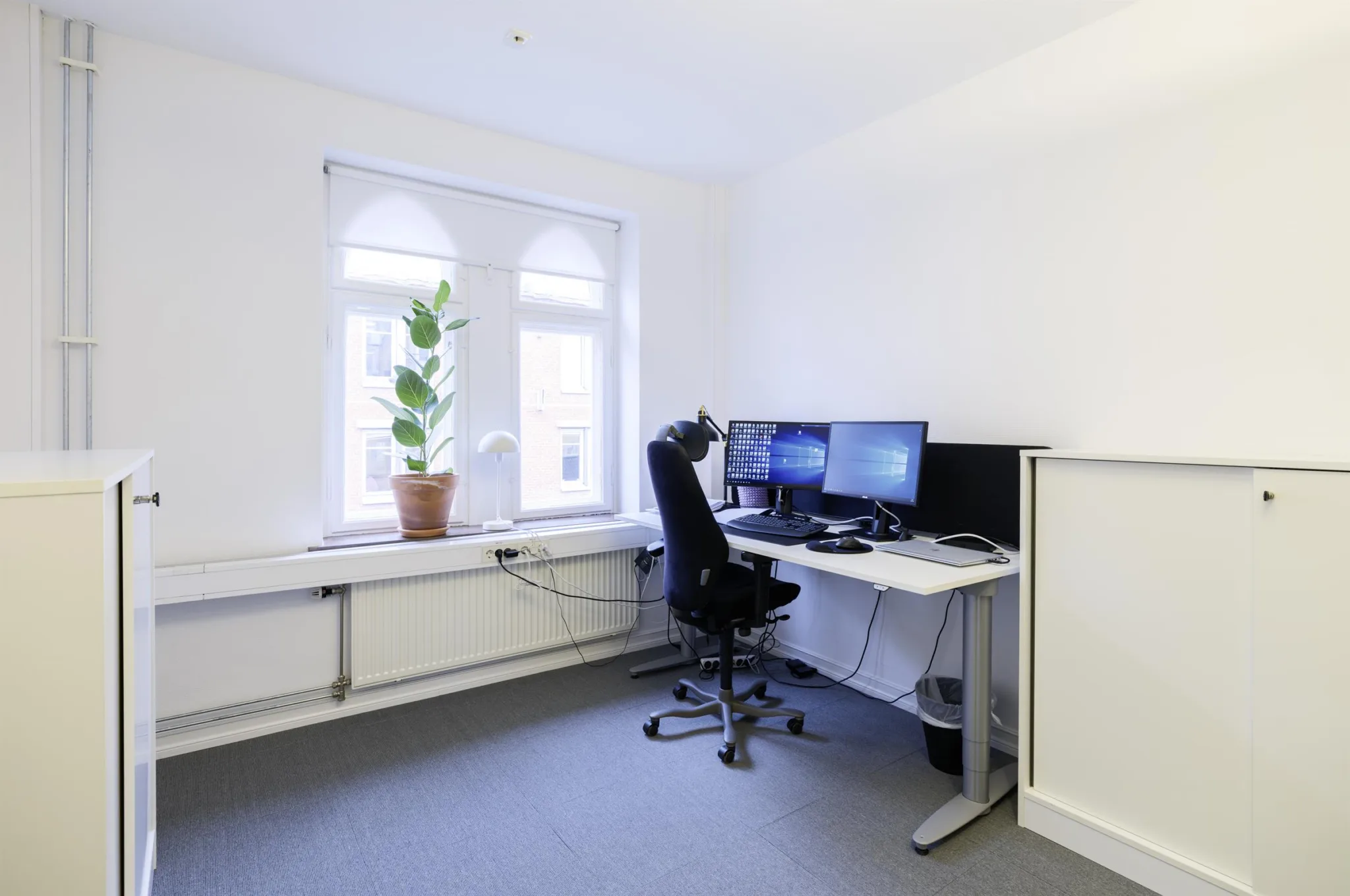2. Ett litet kontor med ett skrivbord, tre skärmar, en kontorsstol och en krukväxt vid ett fönster.