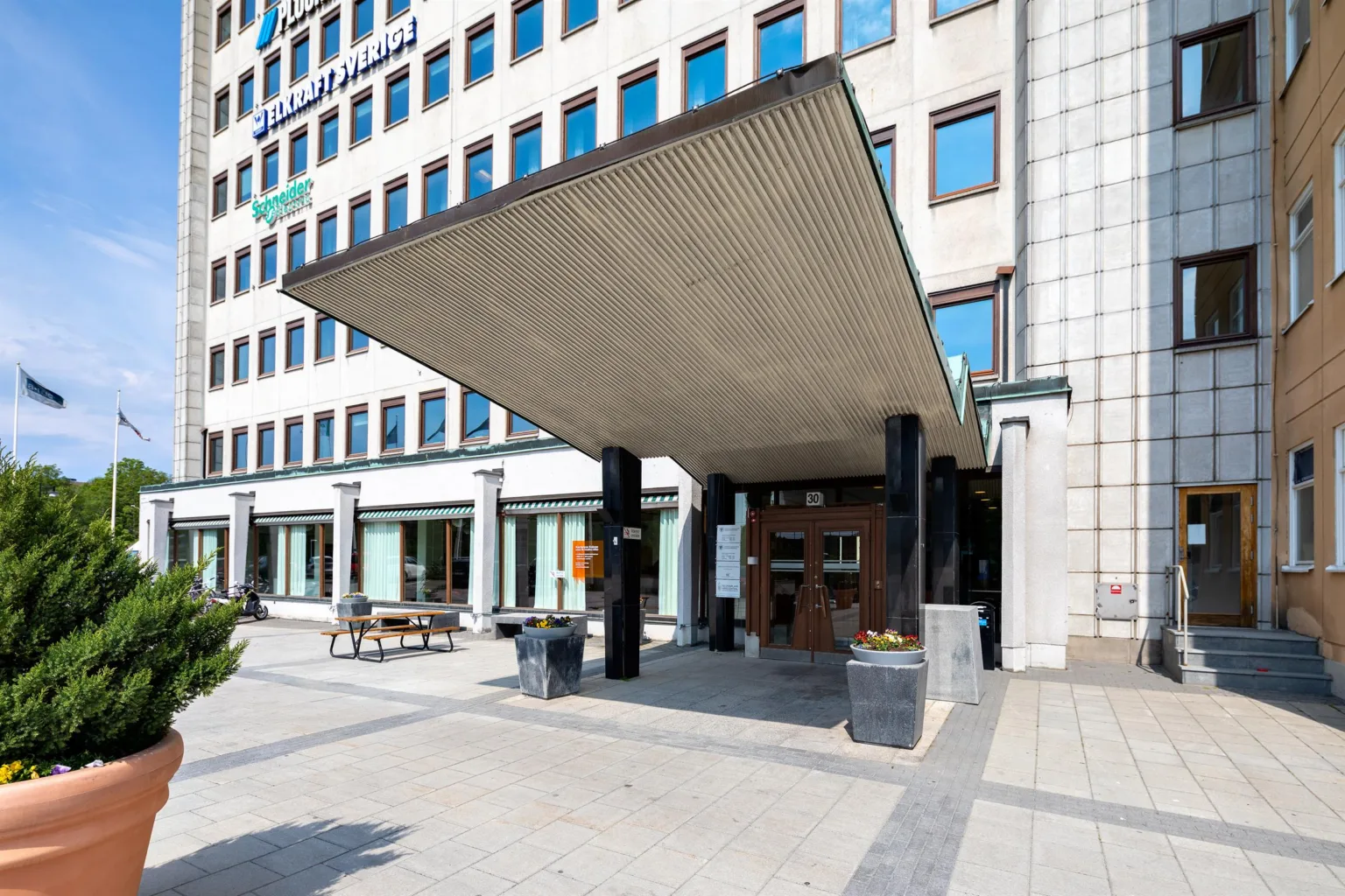 Entré med ett stort tak, glasfönster och pelare framför en modern byggnad.