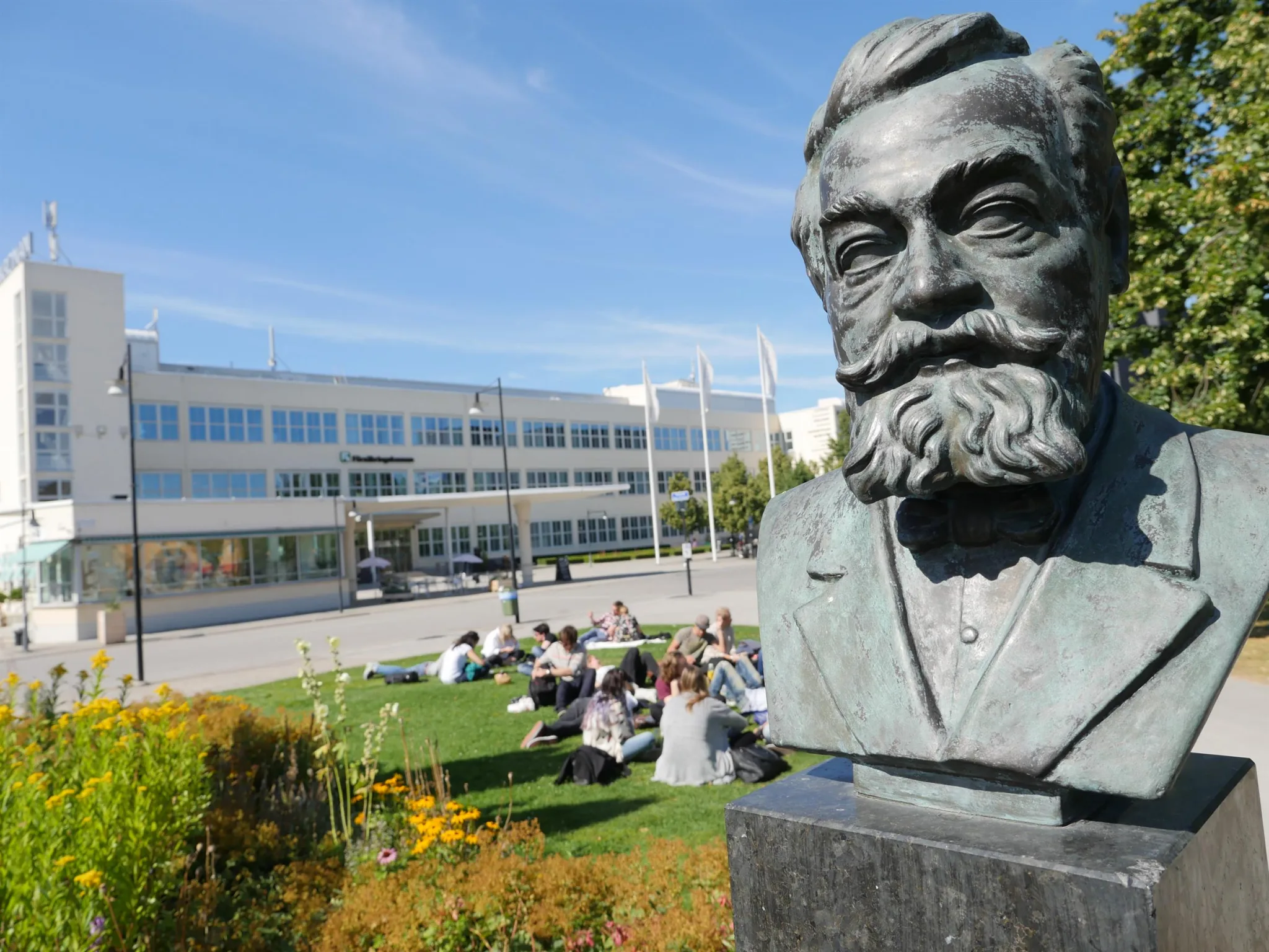 Byst av en man med skägg, grön gräsmatta och människor sittandes utomhus, med en byggnad i bakgrunden under blå himmel.