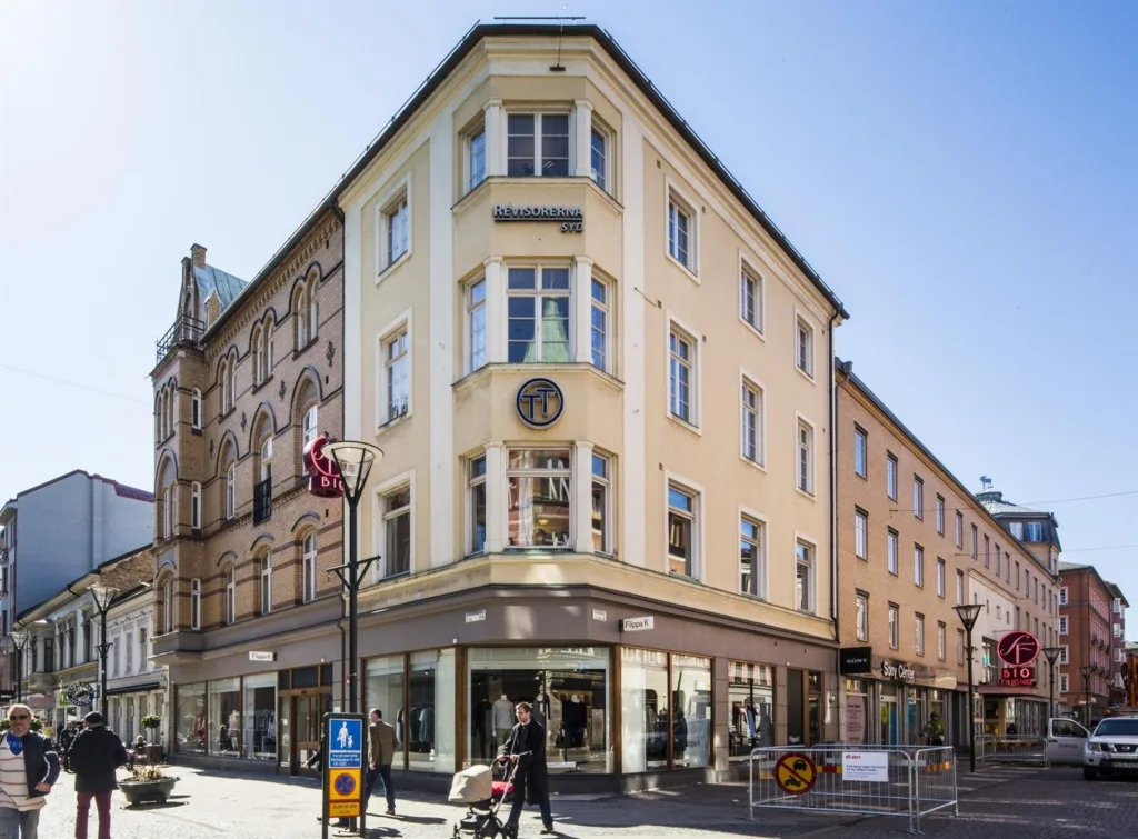 En vit hörnbyggnad med flera våningar, butikslokaler på bottenvåningen och personer som går förbi.