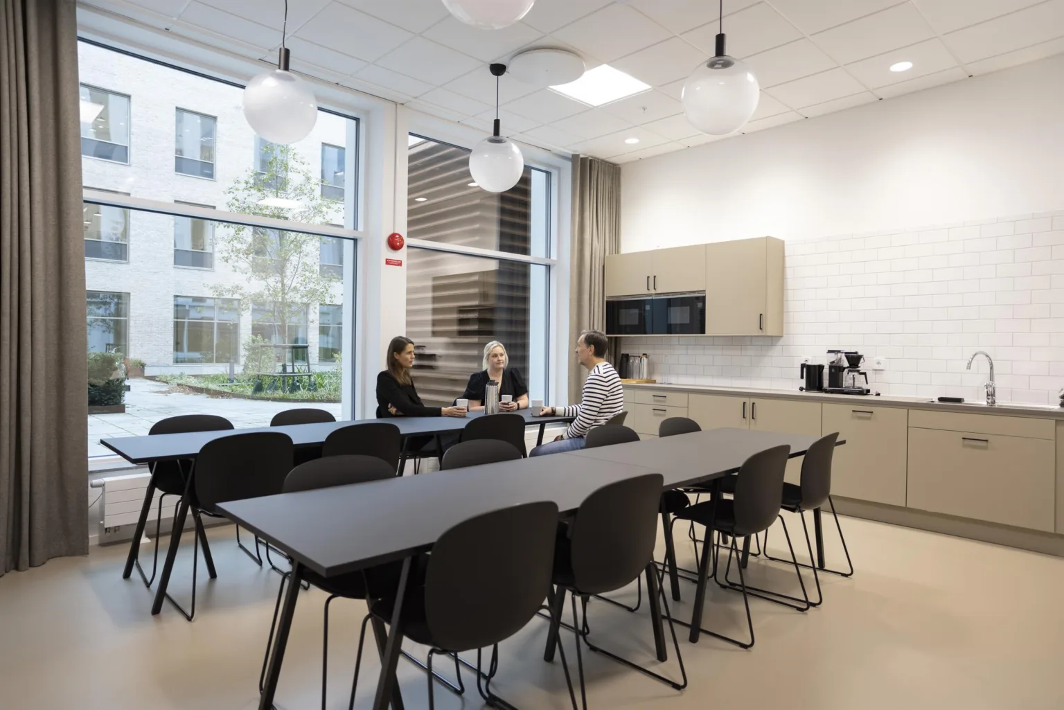 2. Ett modernt personalrum med matbord, stolar, kaffemaskin och stora fönster som släpper in naturligt ljus.