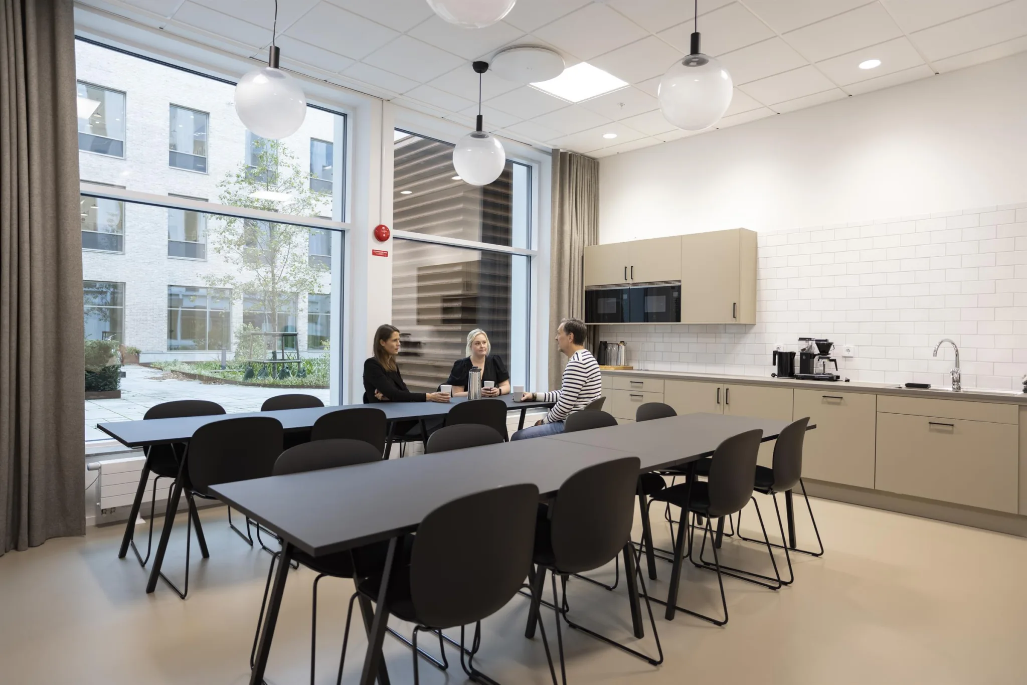 2. Ett modernt personalrum med matbord, stolar, kaffemaskin och stora fönster som släpper in naturligt ljus.